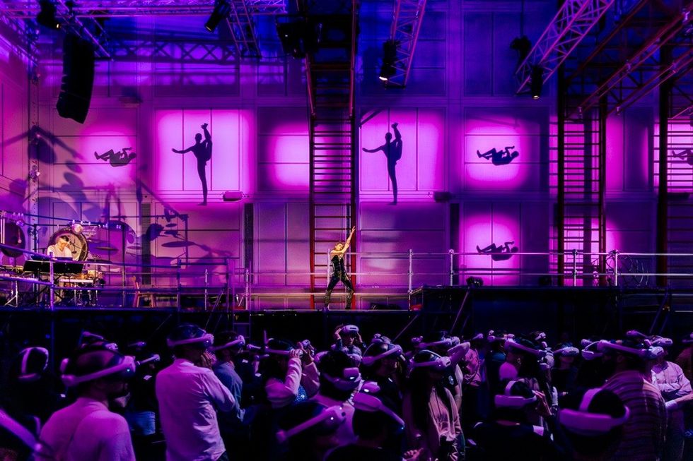Performer on stage with purple lighting, VR audience, and dancing silhouettes on the wall.