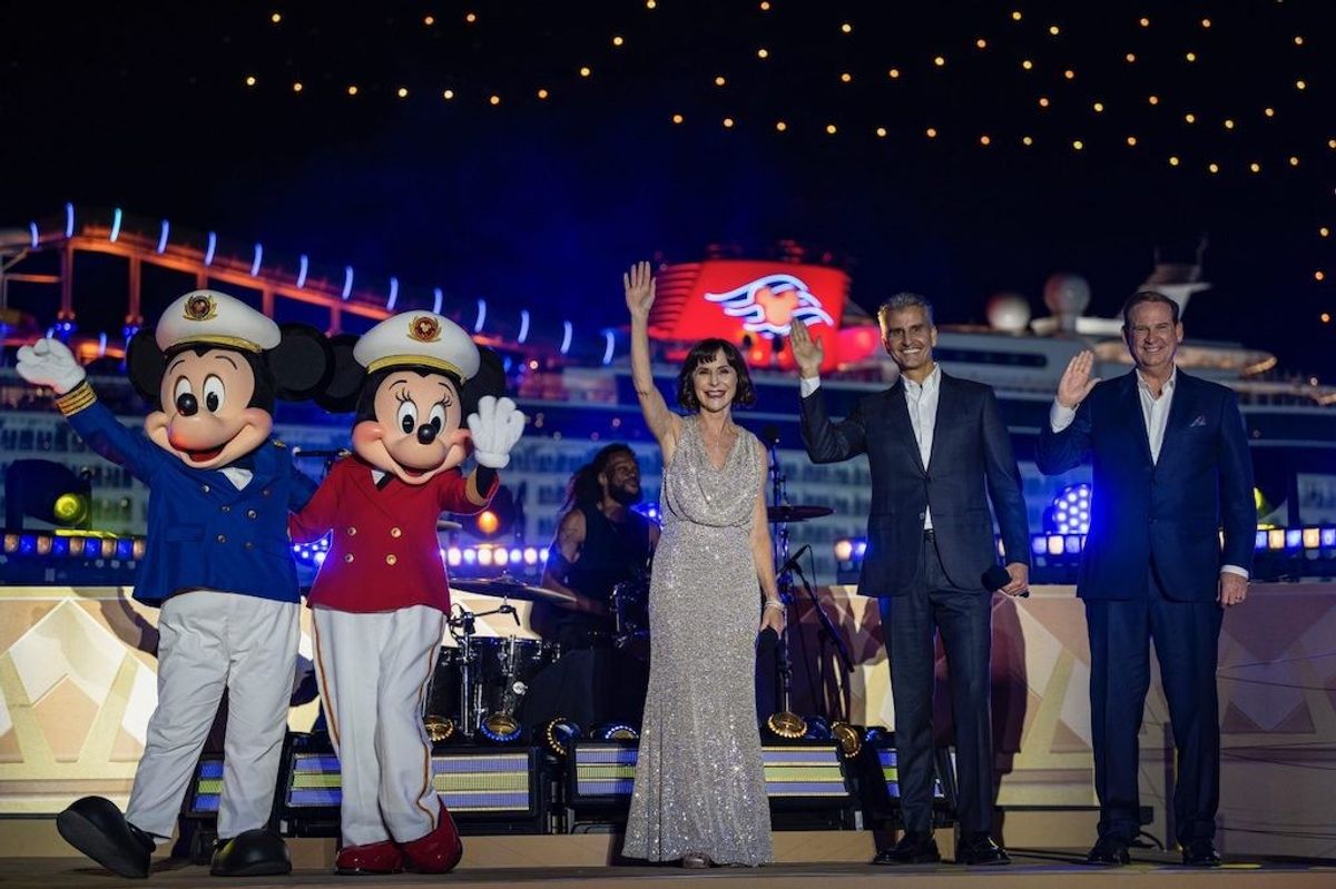 Performers and mascots wave on a brightly lit stage with a ship in the background.