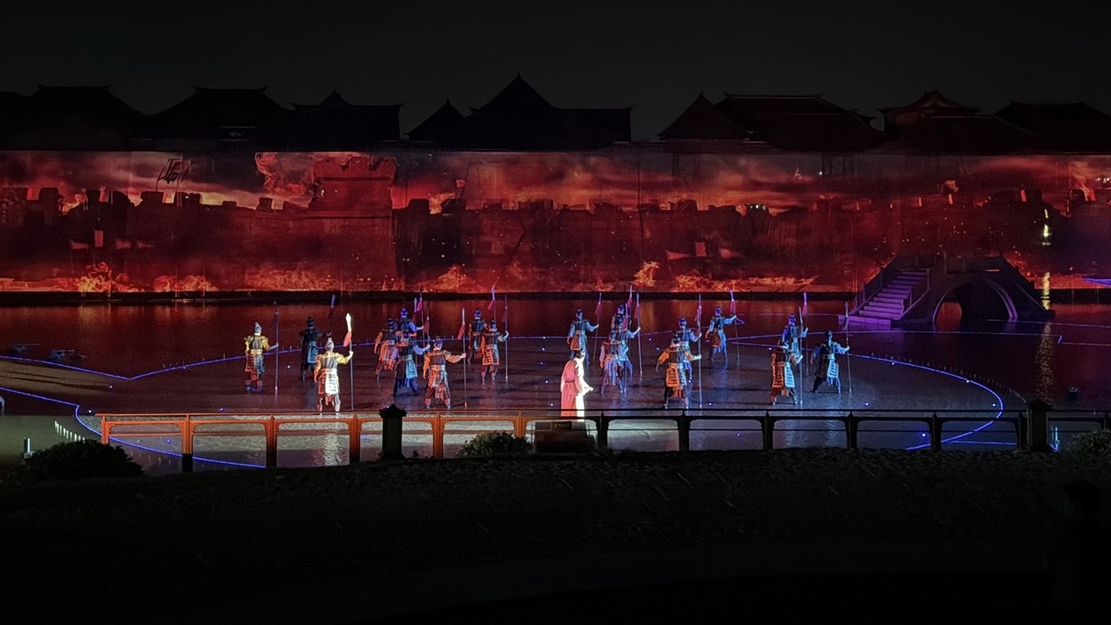 Performers in ancient costumes with a fiery backdrop in a nighttime show titled 'The Wonders of Lu You'