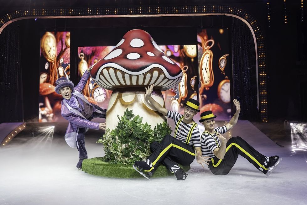 Performers in colorful costumes pose by giant mushroom on stage.