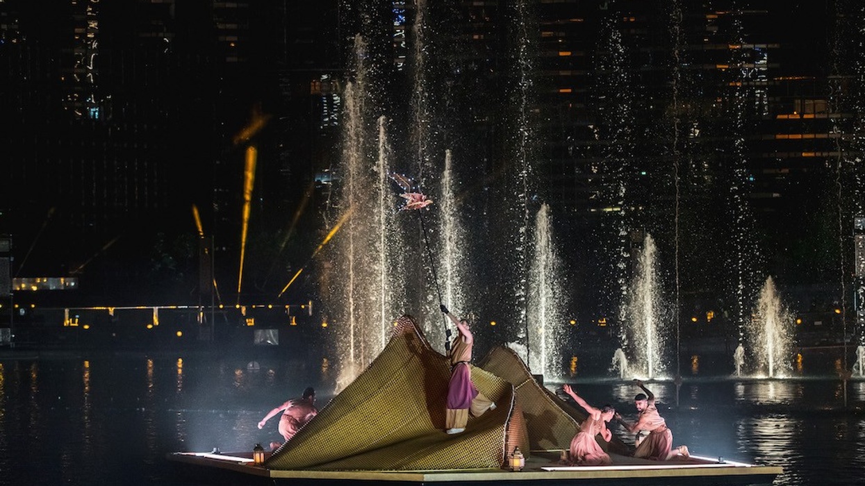 Performers in costumes on a floating stage with water fountains at night.