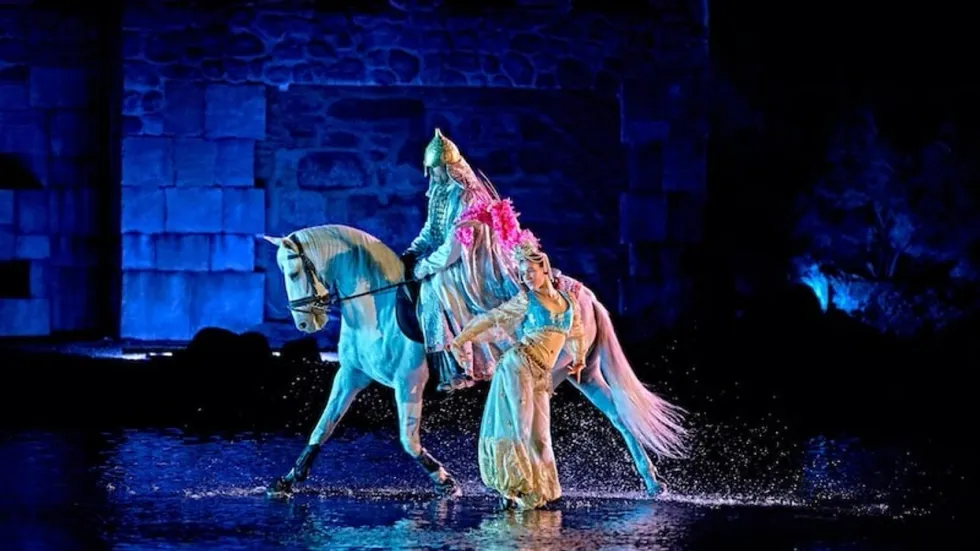 Performers in vibrant costumes on horseback with water splashes in a mystical setting.