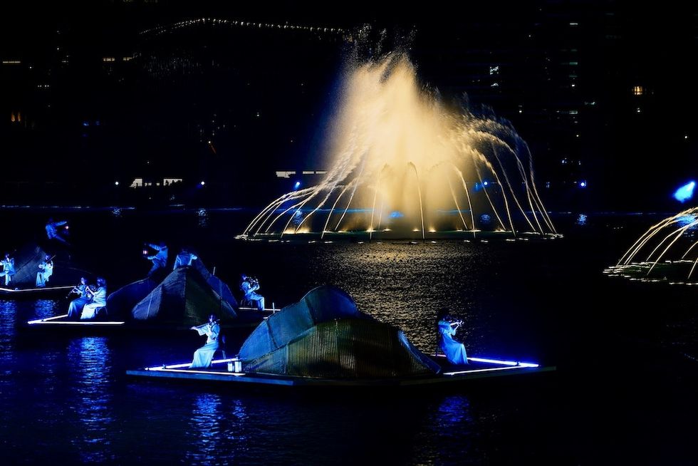 Performers on illuminated boats with a water fountain backdrop at night.