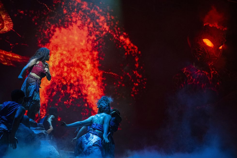 Performers on stage with fiery backdrop and glowing-eyed creature.