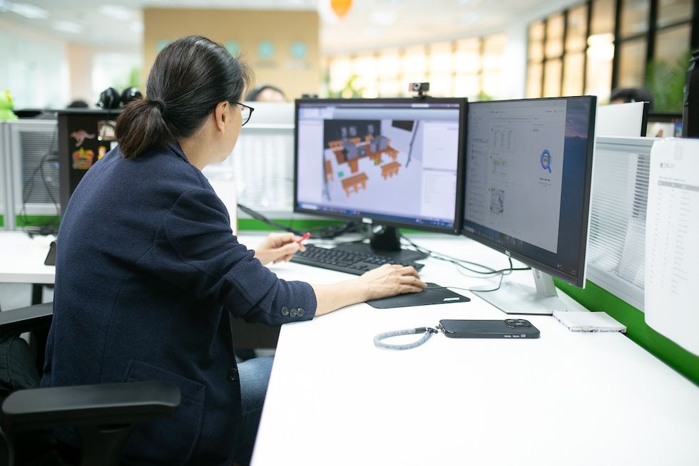 Person at desk in Pico Play's new design space in KL, working on dual monitors, viewing a 3D design model in an office setting.