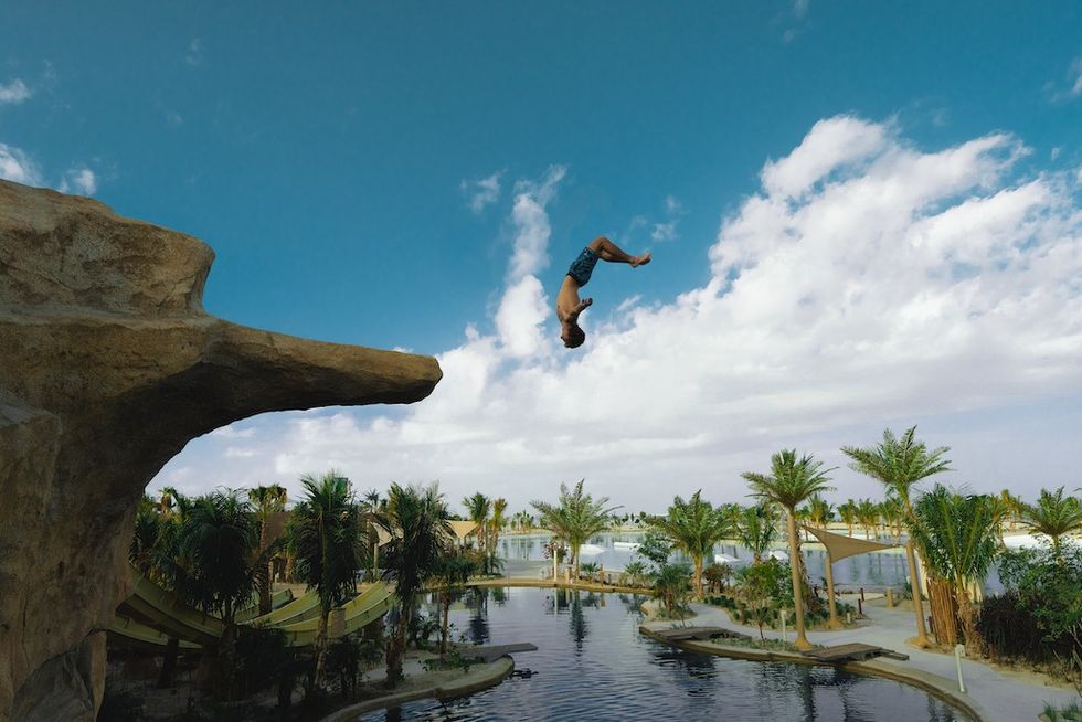 Person flips off cliff into lagoon at Adrena, surrounded by palm trees under a partly cloudy sky.