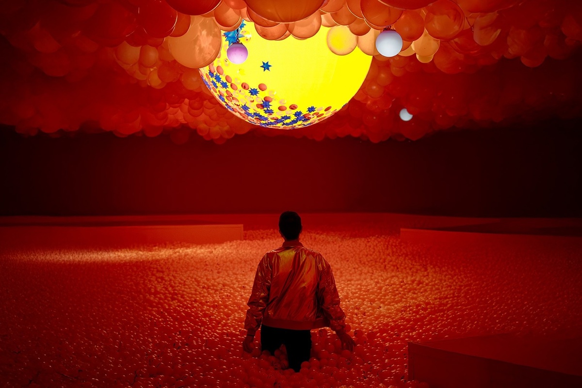 Person in a ball pit under a red-tinted balloon ceiling, with a glowing yellow orb above.