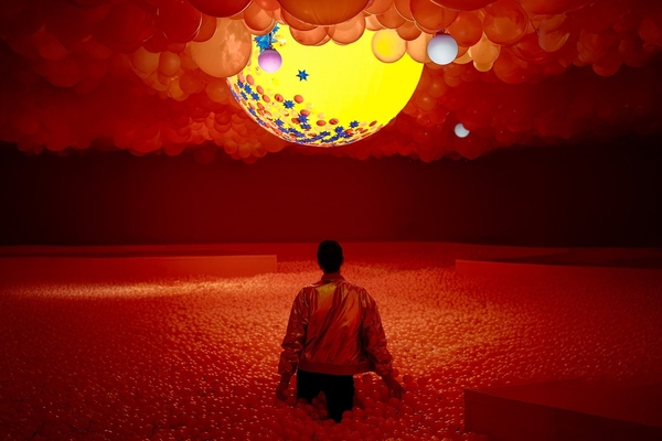 Person in a ball pit under a red-tinted balloon ceiling, with a glowing yellow orb above.