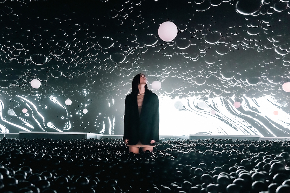 Person in a dark room with a swirling light pattern, surrounded by black balls and hanging spheres.