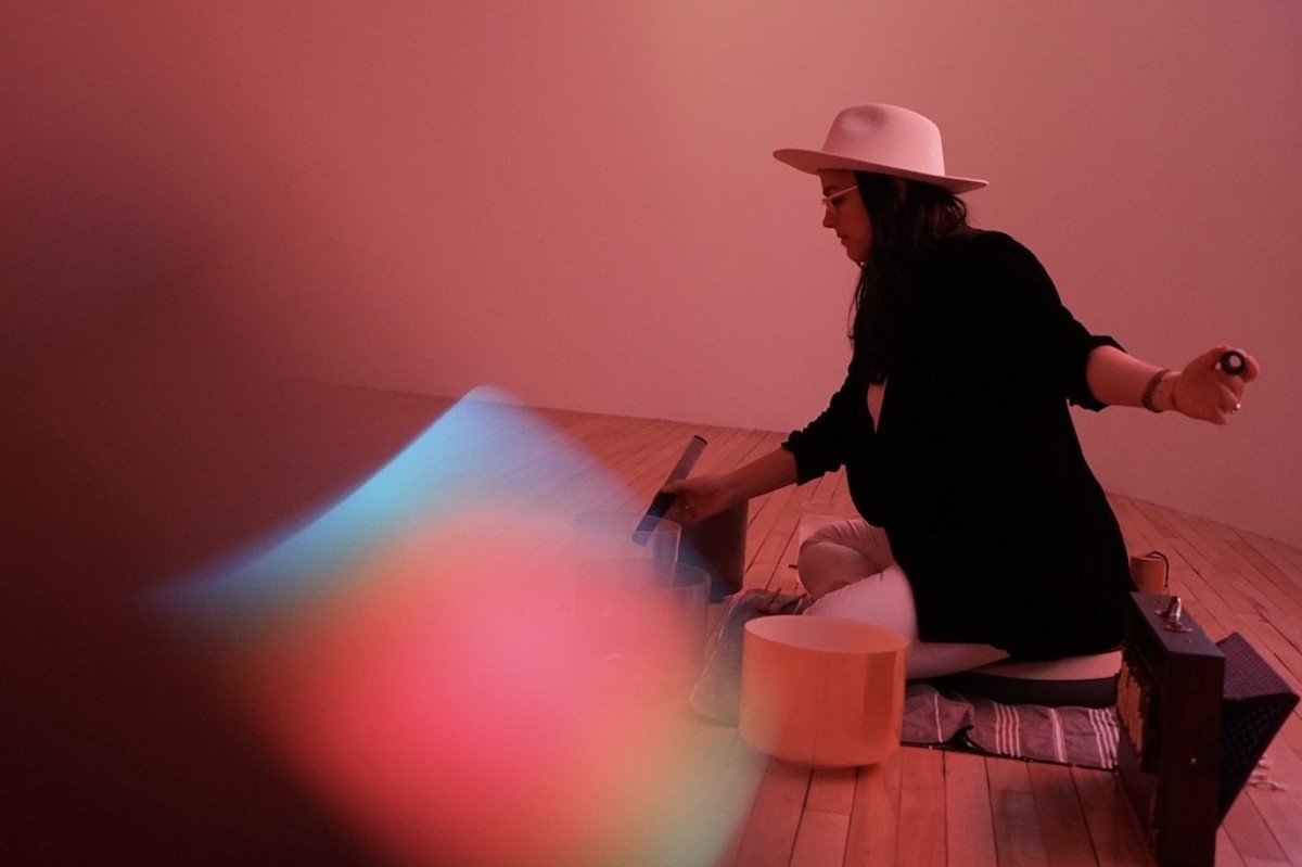 Person in a hat playing crystal singing bowls, surrounded by soft red lighting.