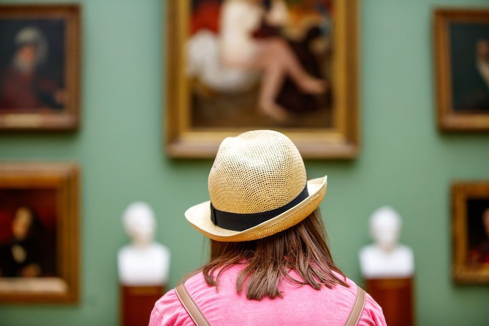 Person in a hat viewing paintings and busts in an art gallery.