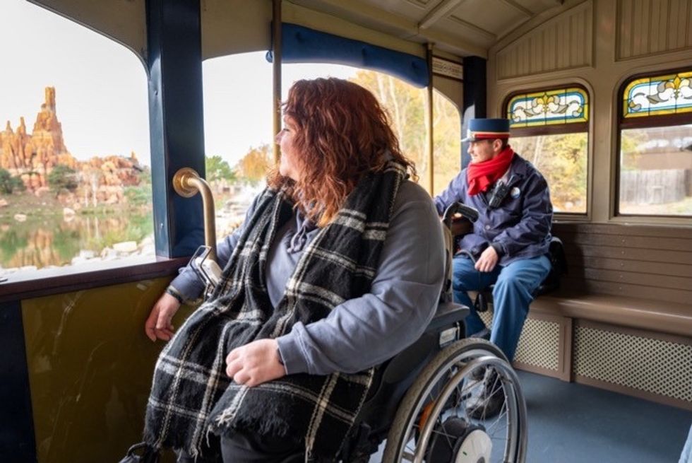 Person in a wheelchair enjoying The Disneyland Railroad with scenic rocky view, conductor nearby.