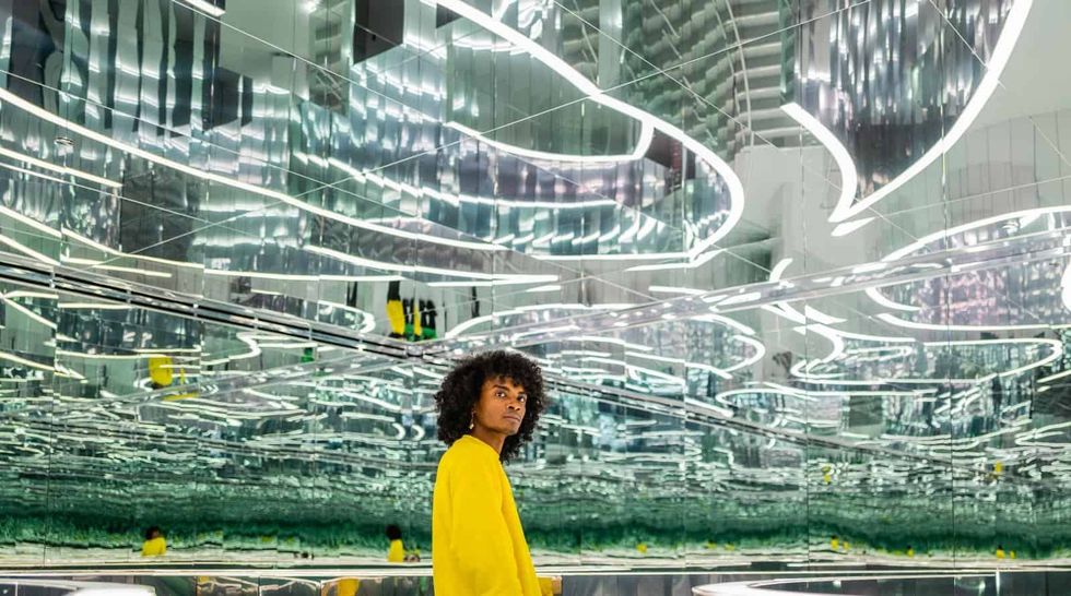 Person in a yellow sweater in a mirrored, reflective room with bright, abstract lights at Superblue