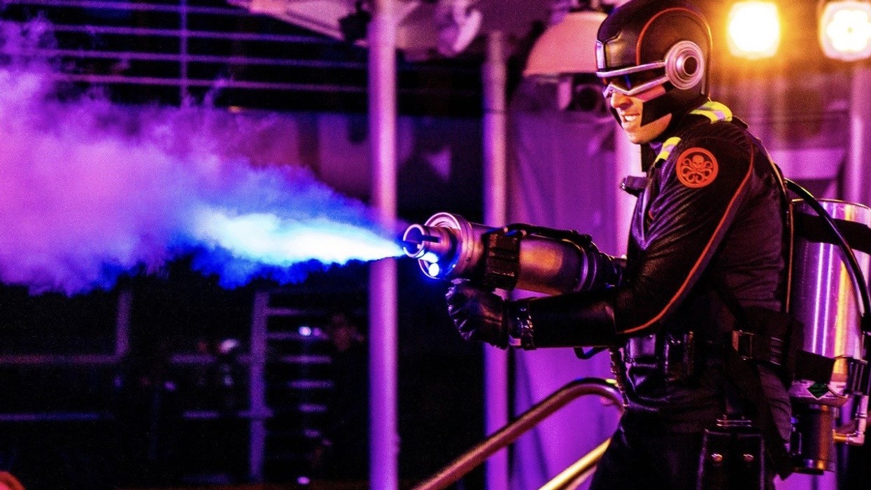 Person in futuristic costume spraying colored smoke from a device, under purple lighting.