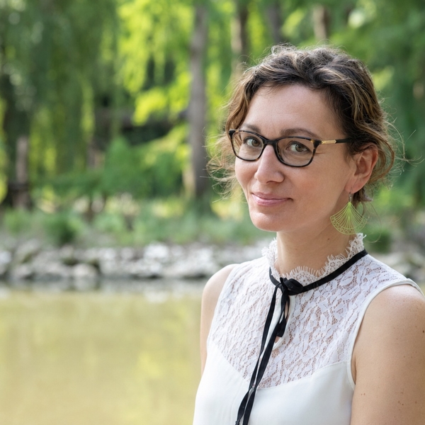 Person in glasses and a white top smiling outdoors with greenery and water in the background.