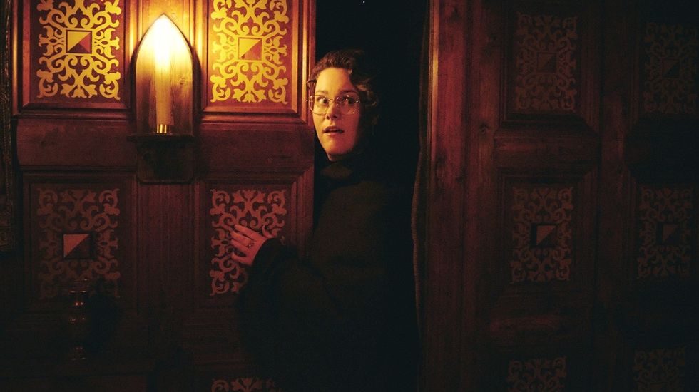 Person in glasses stands by ornate wooden door under warm lighting.