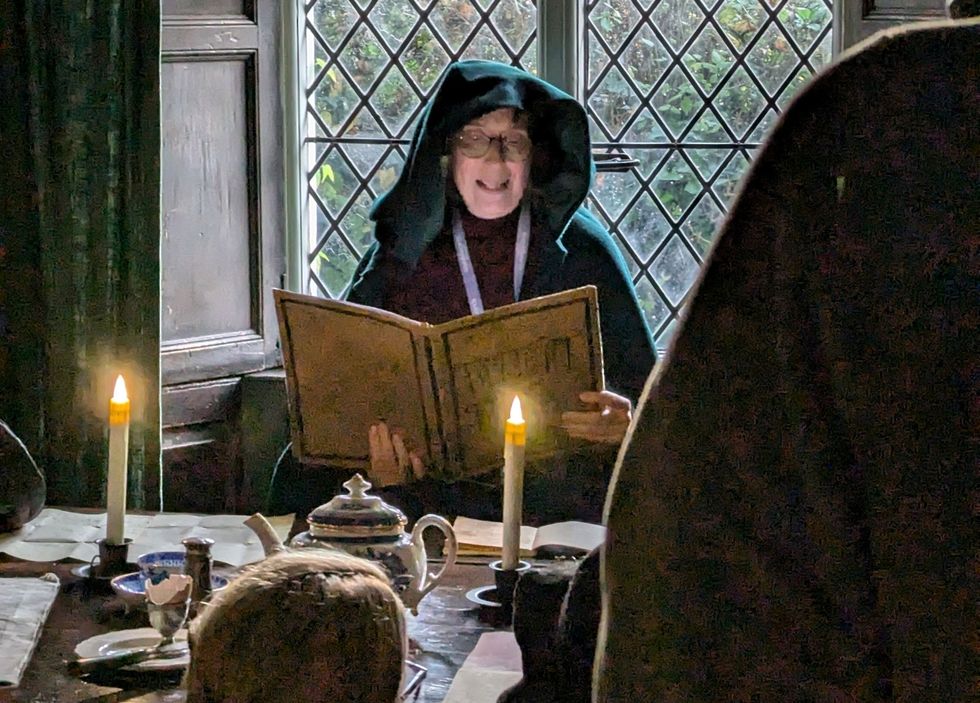 Person in hooded cloak reads a large book by candlelight in a dim room.