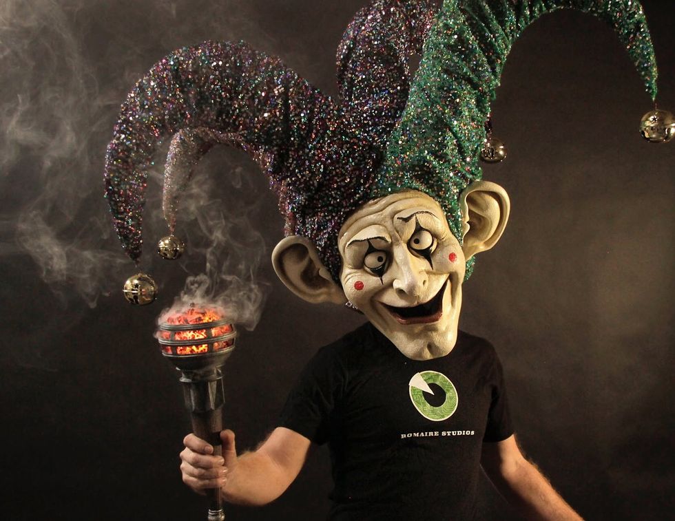 Person in jester mask holding smoking torch, wearing sparkly hat with bells.