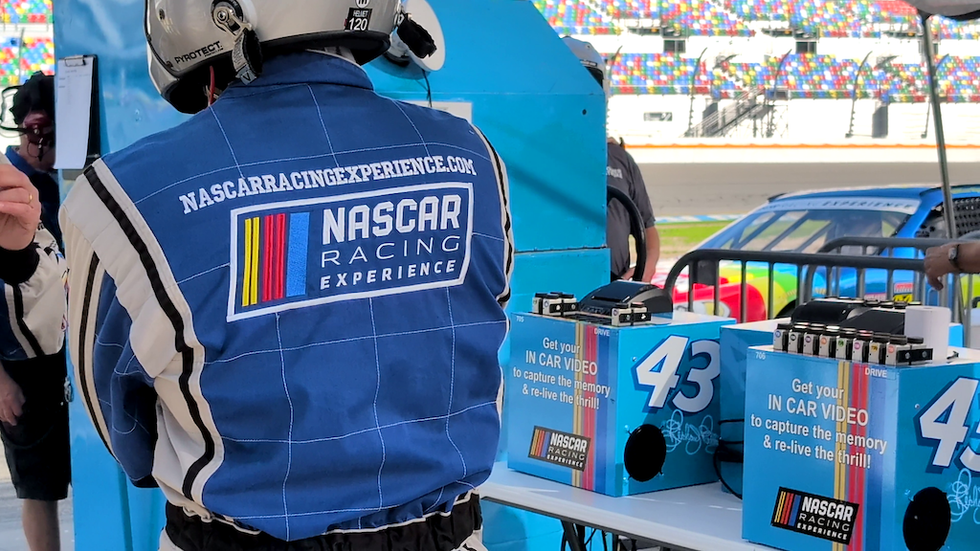 Person in NASCAR jumpsuit near in-car video displays at a racetrack.