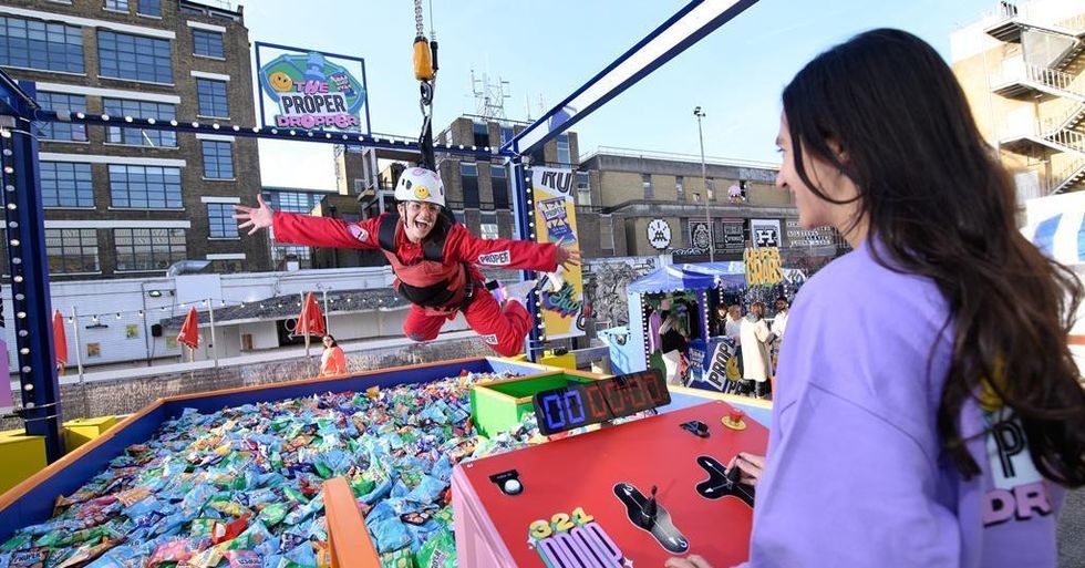 Person in red gear suspended above a giant claw machine filled with snack bags.