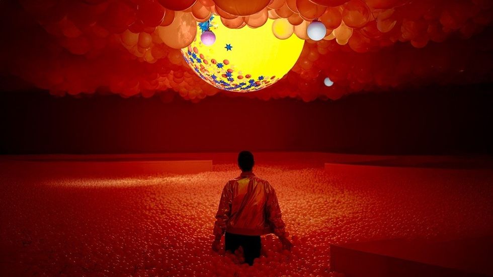 Person in red-lit ball pit gazes at a large, colorful balloon installation.