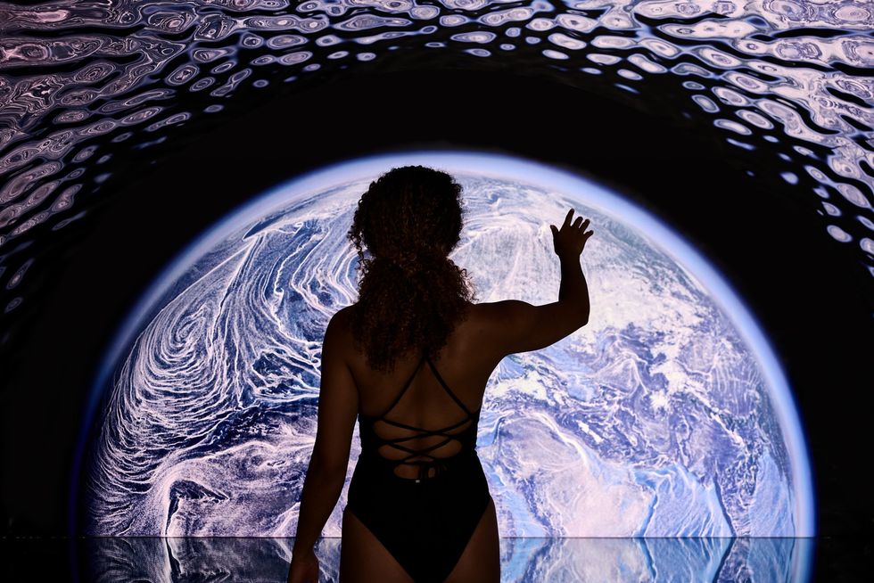 Person in swimsuit reaches towards large Earth-like display, wavy patterns overhead.