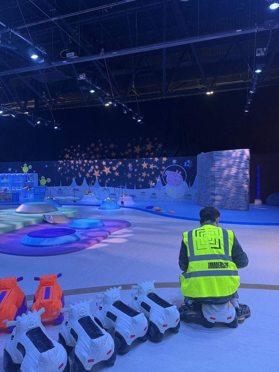 Person in yellow vest at a colorful indoor play area, Peppa Pig's Space Adventure, with toy cars and starry decorations.