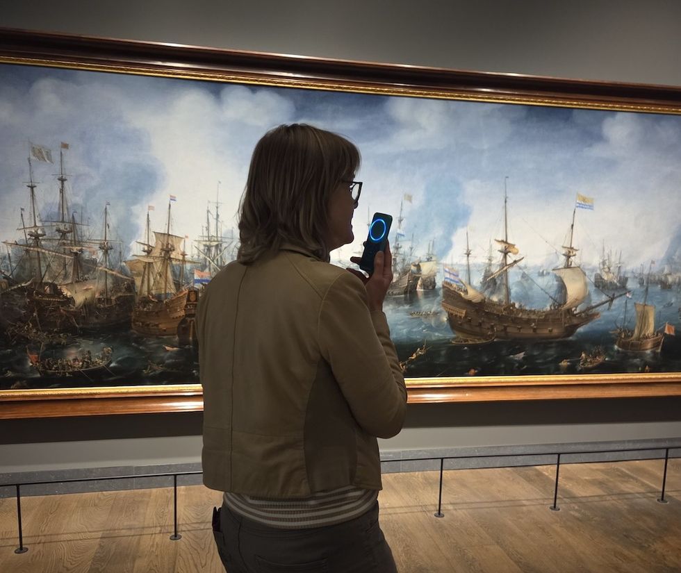 Person listening to an audio tour in front of a large maritime painting in a gallery.