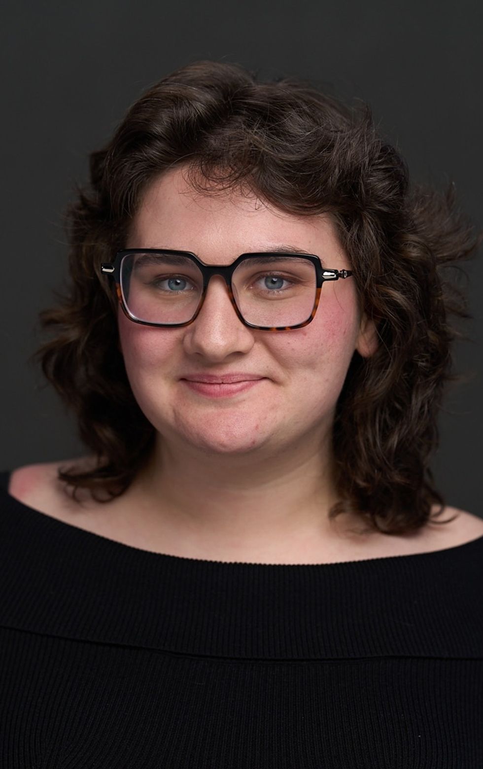 Person named Madeline Miller, with curly hair and glasses, wearing a black top, against a dark background.