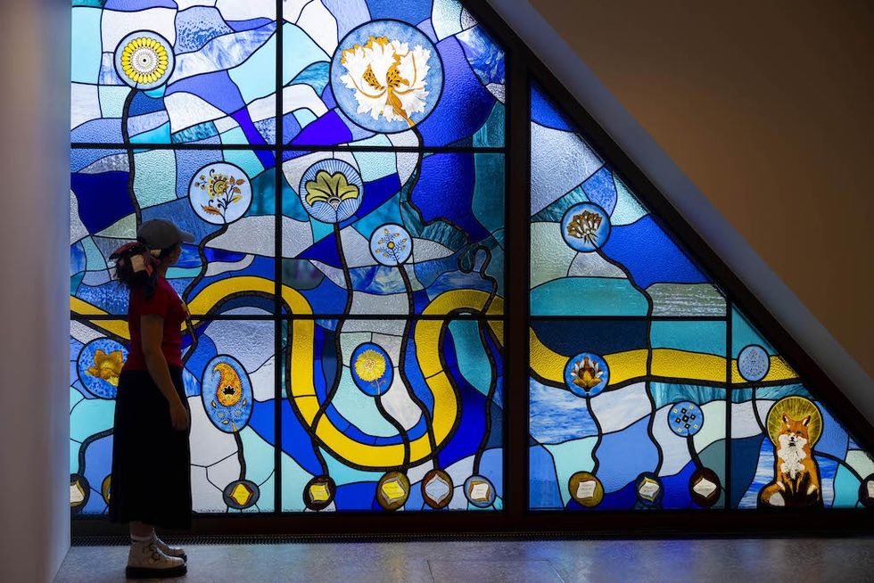 Person observing Towards A Civic Museum\u2019 by Tania Bruguera, a vibrant blue and yellow stained glass mural, at V&A East