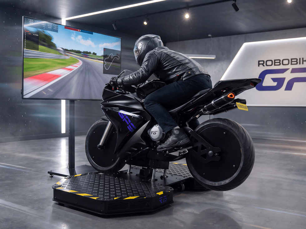 Person on Robobike motorcycle simulator in a modern room with large screen track display.