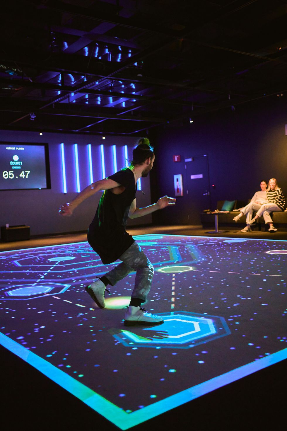 Person playing interactive floor game with neon graphics, two people seated in the background.