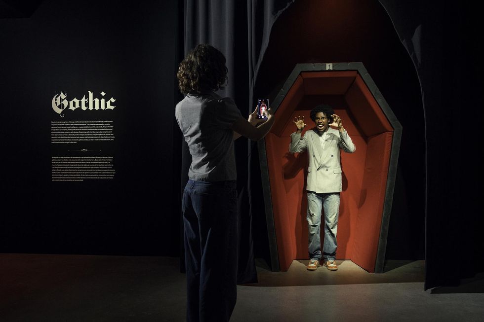 Person poses in a coffin, another takes a photo; "Gothic" text on wall at the Academy Museum