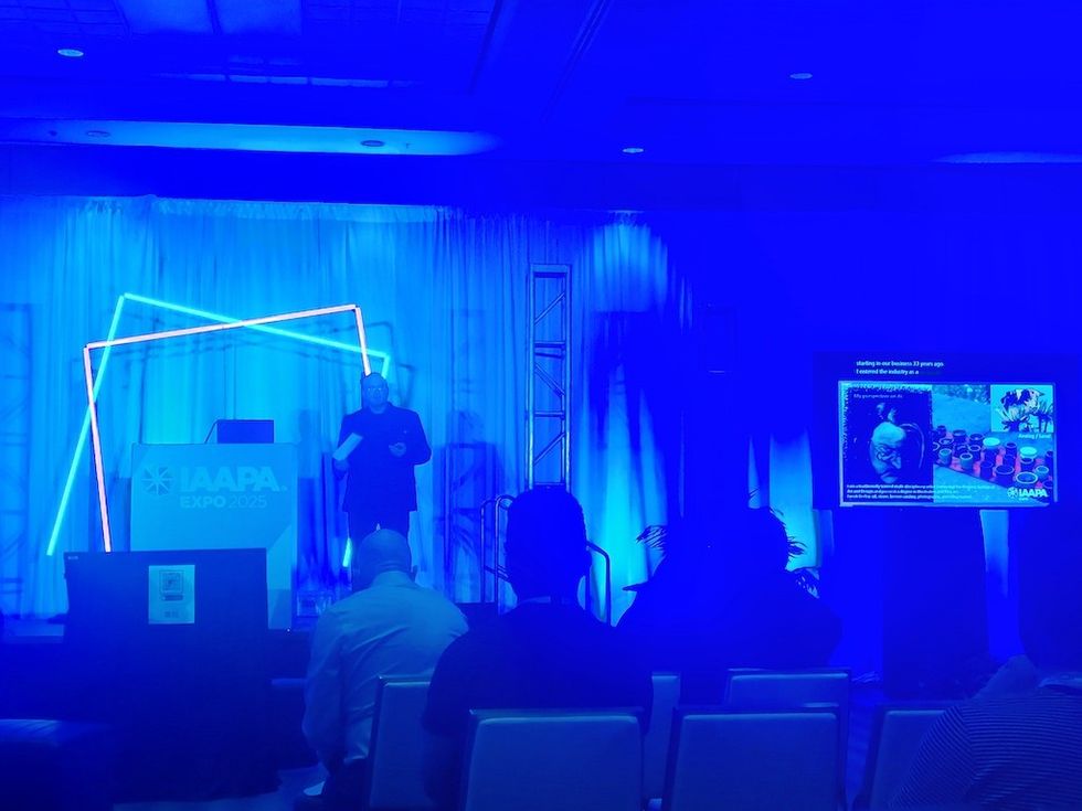 Person presenting at IAAPA Expo 2025 with a blue-lit stage and screen on the right.