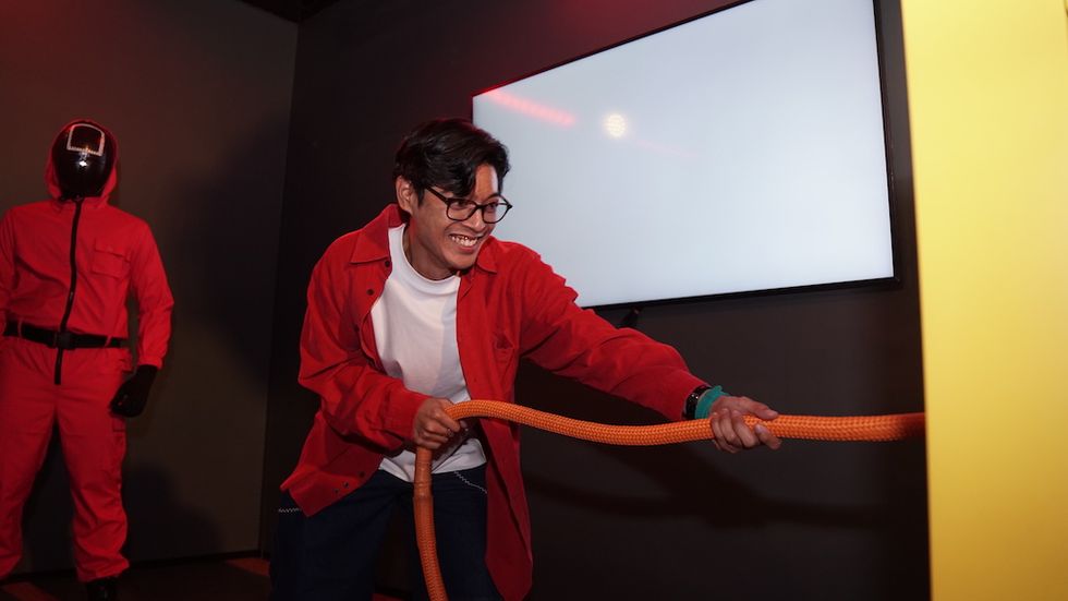 Person pulling rope with smiling expression; hooded figure in pink outfit watches nearby.