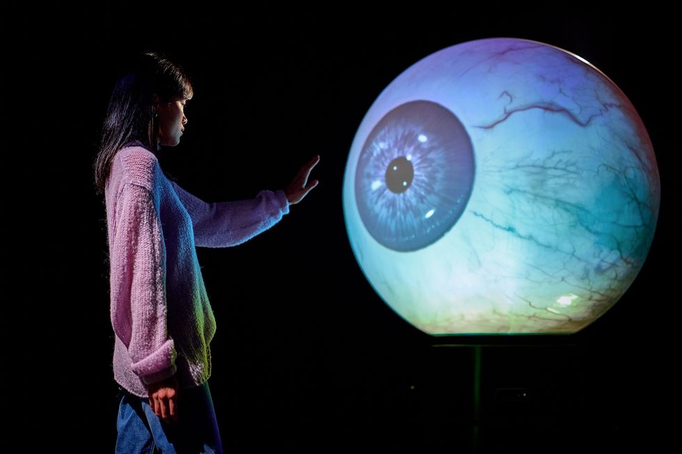 Person reaching towards a large, illuminated spherical eye in a dark room at Prototype: The Experimental Museum