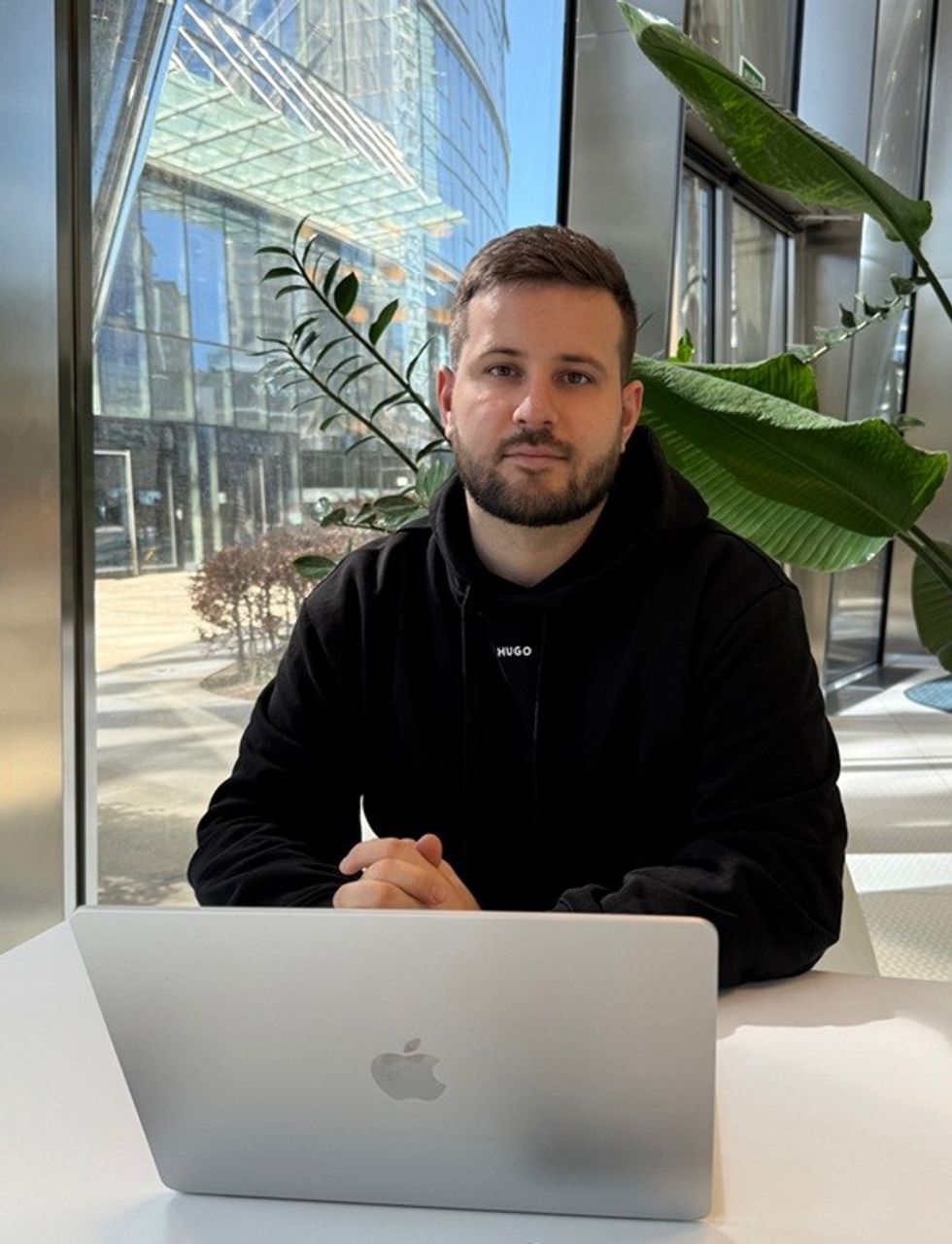 Person sitting at a laptop in a modern building with large windows and tropical plants.