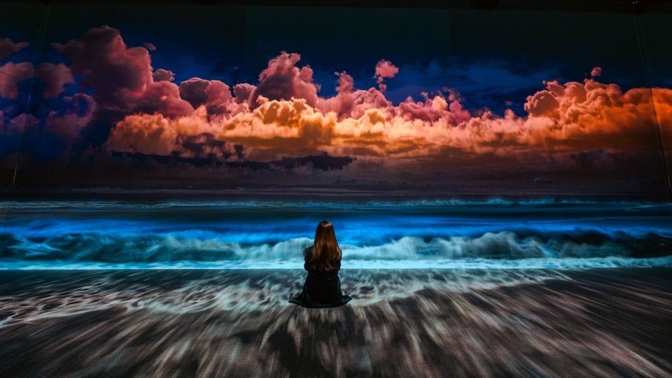 Person sitting on a beach watching vibrant clouds and waves at sunset.