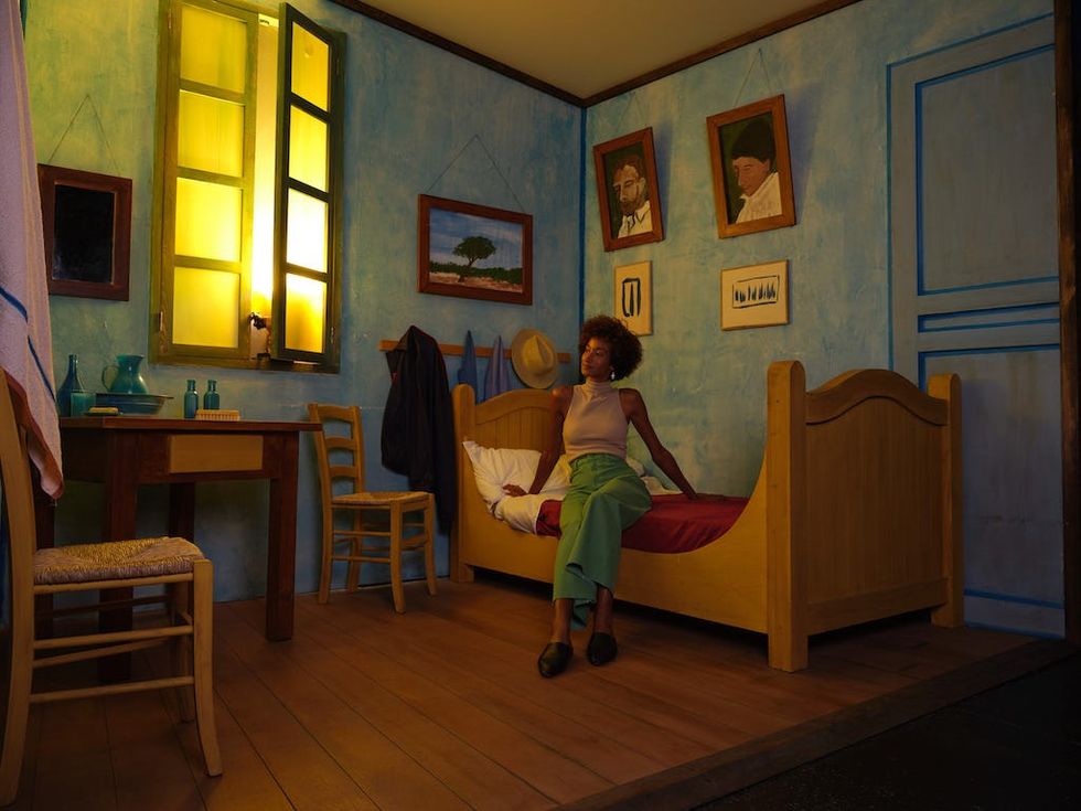 Person sitting on a bed in a Van Gogh-inspired room with paintings and a glowing window.