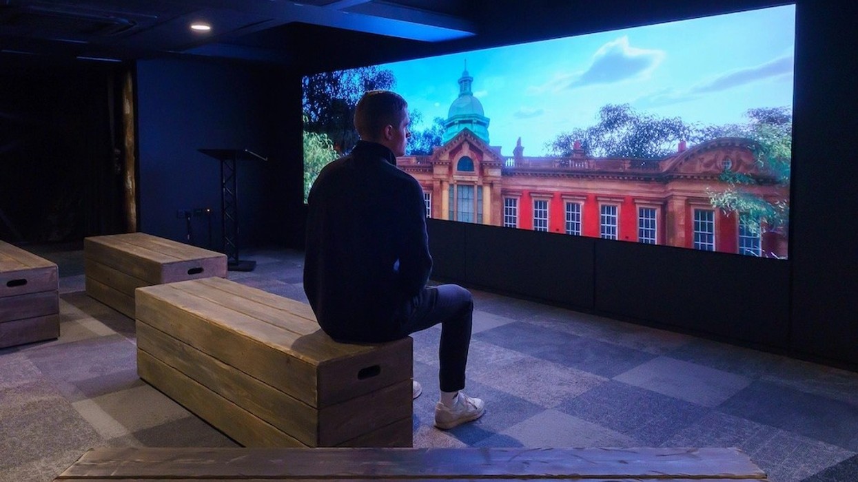 Person sitting on a bench at Redhills Durham Miners Hall, watching a large screen displaying a building with trees.