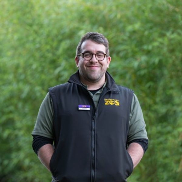 Person smiling, wearing a zoo vest, stands in front of lush greenery.