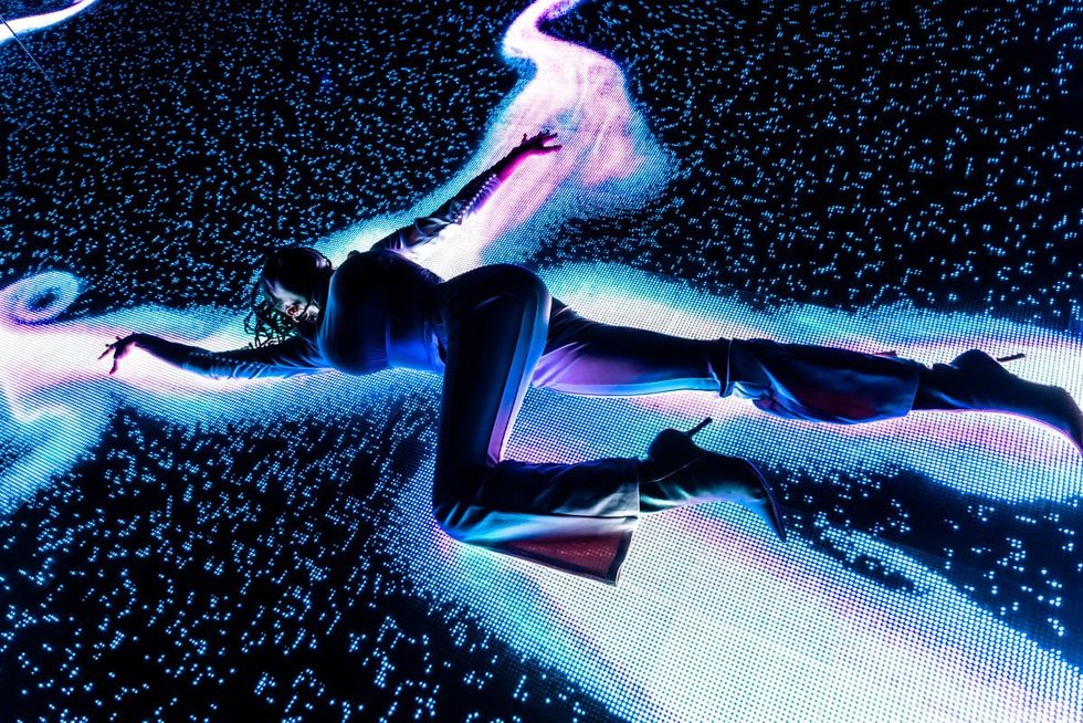 Person sprawled on illuminated digital floor with abstract blue and purple light patterns at WNDR Museum