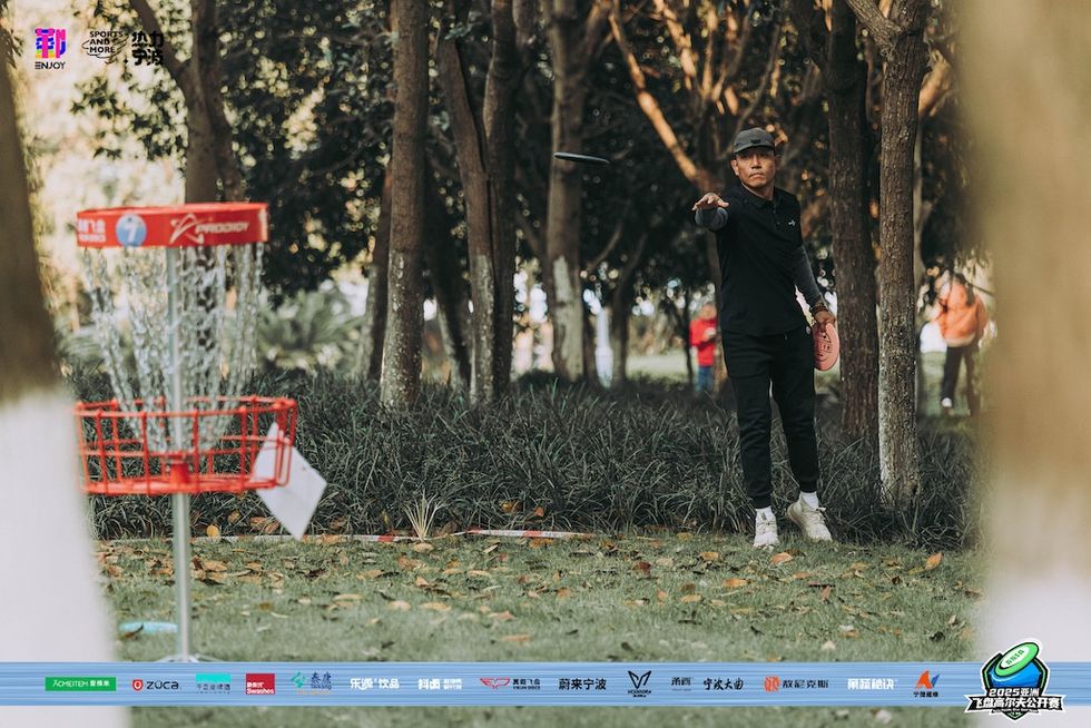 Person throwing a disc in a disc golf game, aiming for the basket in a wooded area.