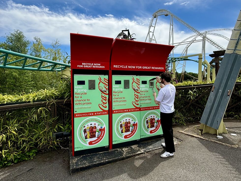 Person using a Coca-Cola recycling station near a roller coaster on a sunny day.