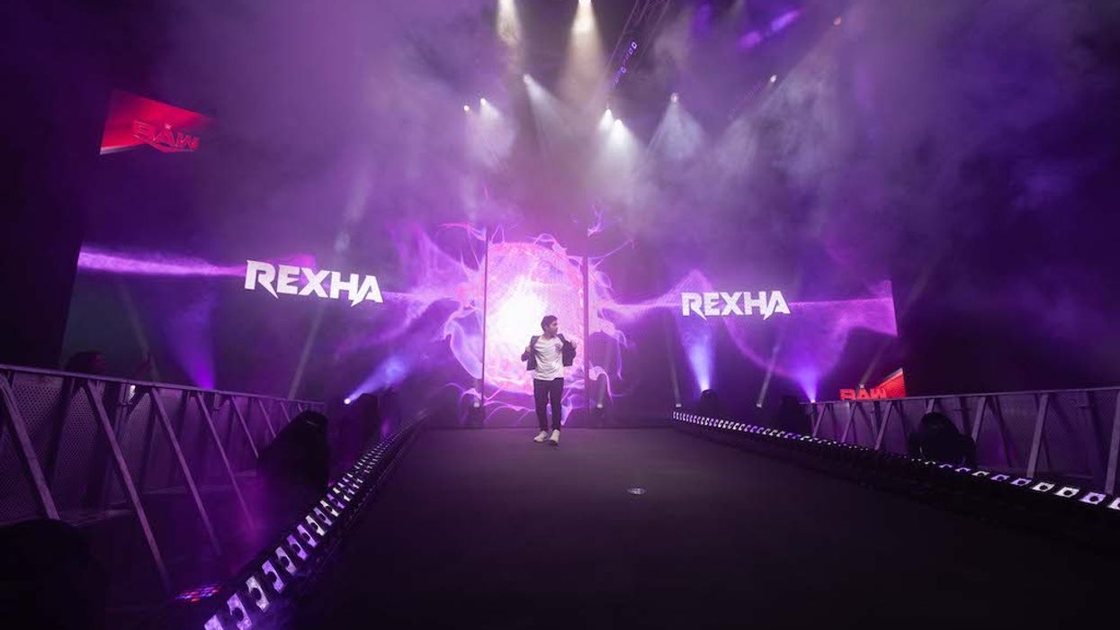 Person walking on stage with purple lights and "REXHA" in the background.