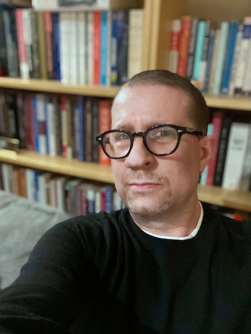 Person with glasses, Dr Josh Perelman, in front of a bookshelf, wearing a black sweater.