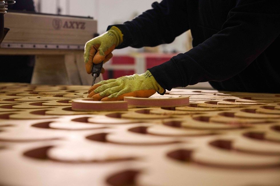 Person with gloves cutting circular shapes from a wooden sheet using a tool.