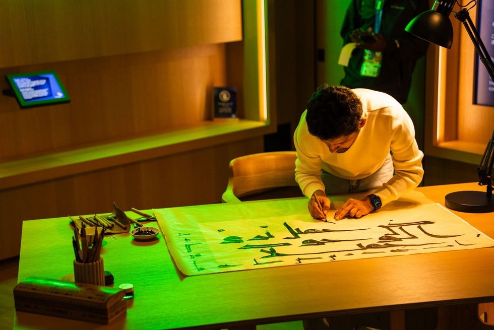 Person writing Arabic calligraphy on a large sheet at a well-lit wooden desk.