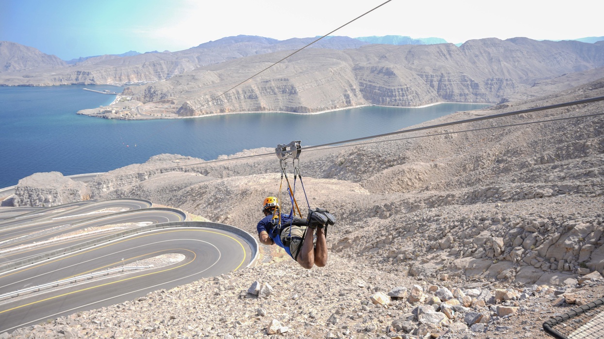 Person ziplining over rocky mountains, curving road, and blue sea below.