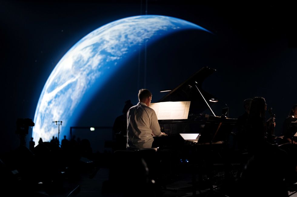 Pianist performing with Earth projected in the background, dark concert setting.